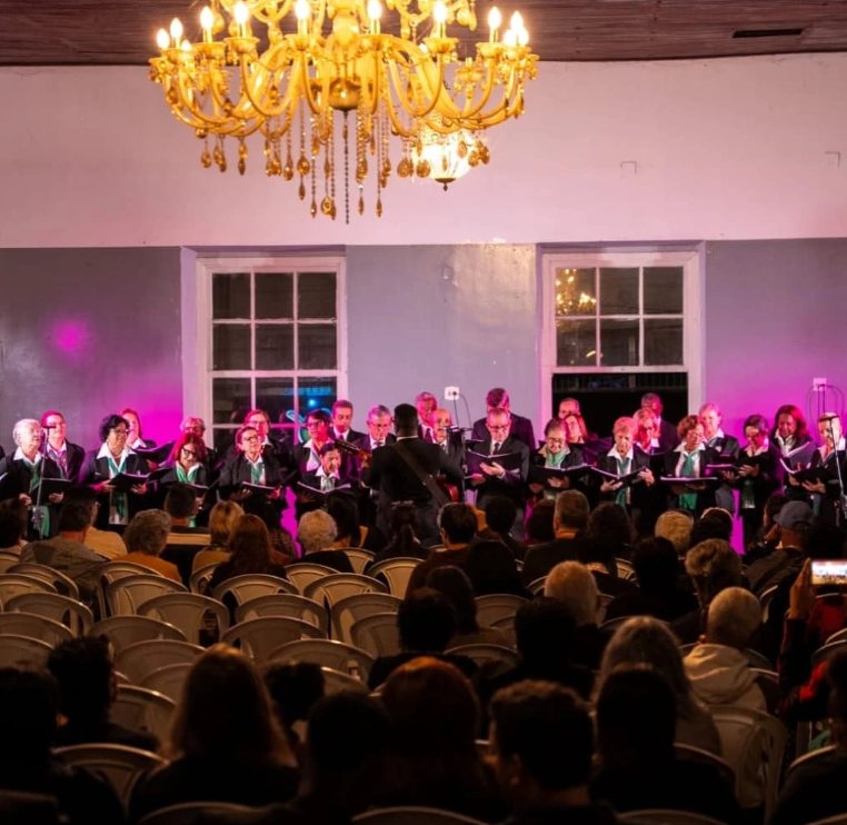 Coral se apresentando sob um lustre em salão histórico, diante de plateia lotada, durante o Encontro Brasilidades