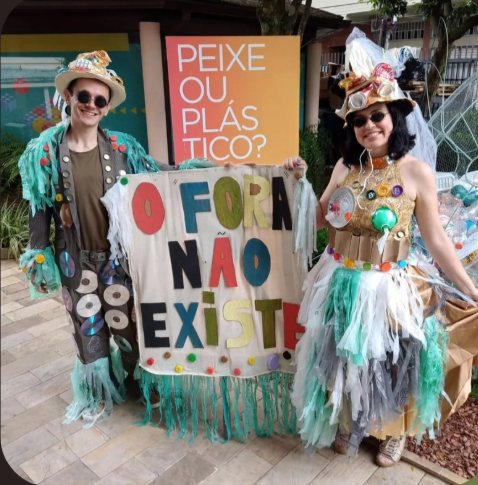 Dupla de artistas em figurinos feitos de material reciclado segurando a faixa O Fora Não Existe, ao lado do painel Peixe ou Plástico?