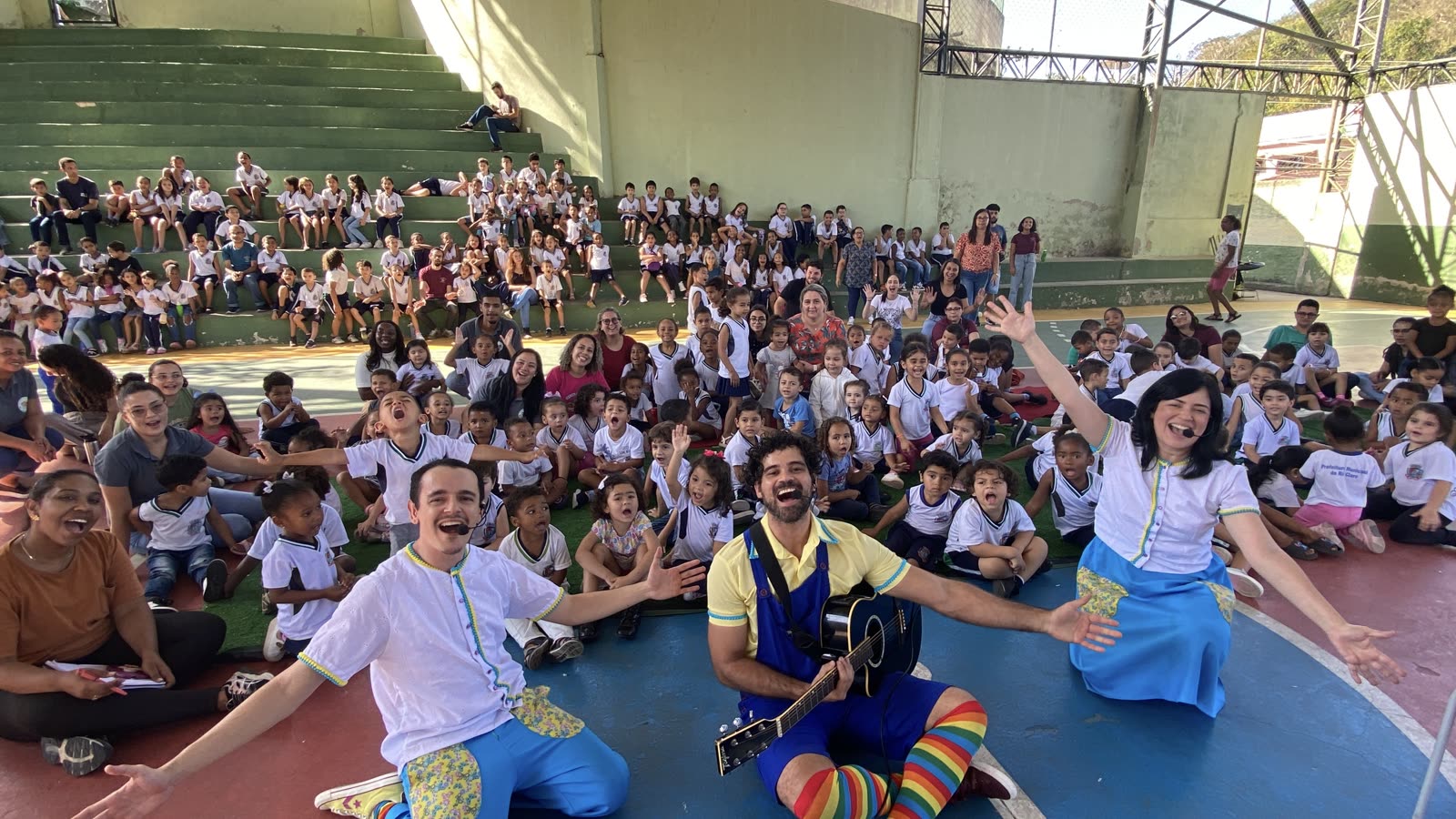 Elenco da Valiante ao final de uma apresentação em quadra escolar, cercado por centenas de crianças e educadores