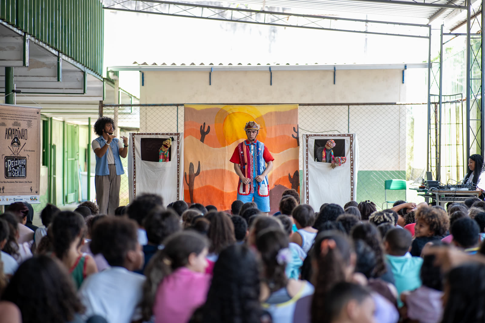 Apresentação do espetáculo O Reizinho do Sertão em pátio escolar, com ator, mamulengos e intérprete de Libras diante de uma plateia lotada de crianças